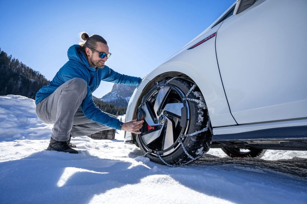 A man in a blue jacket fitting a König Reflex Edge snow chain on the front tyre of a white Volkswagen Golf GTI in a snow-covered mountain parking area on a sunny winter day.