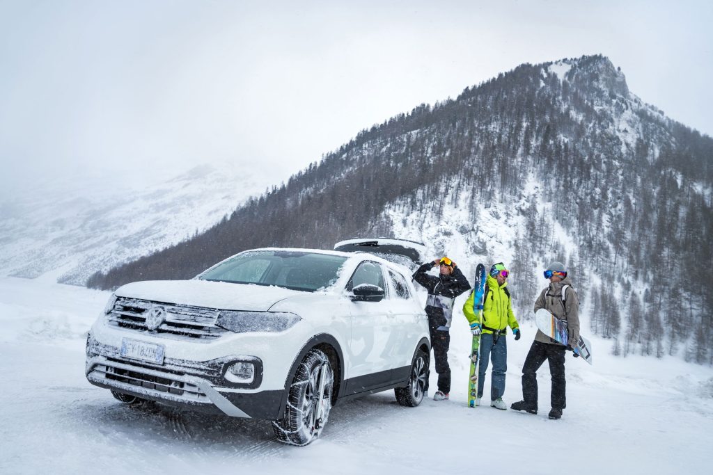 A group of three men are talking near a white Volkswagen T-Cross fitted with König Easy-fit CU-9 snow chains in a snow-covered mountain parking area during a snowfall.