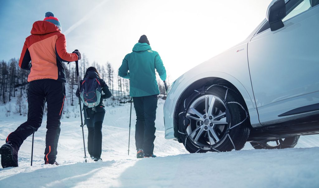 A group of three people walking in the snow with walking poles near a white Audi Q3 fitted with König Reflex Cross snow chains, with snow-covered mountains in the background during a sunny winter day.