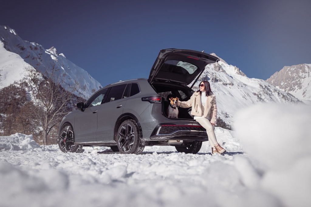 A woman sitting on the edge of the open trunk of a Volkswagen T-Roc with her dog inside, the car fitted with König K-Summit XL snow chains in a snowy mountain area.
