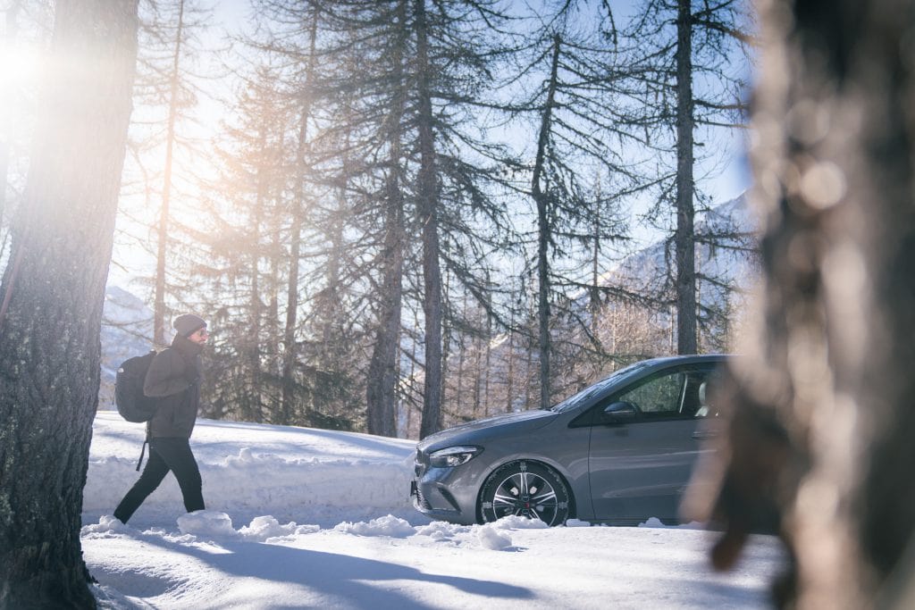 A man with a black backpack is walking towards a grey Mercedes B-Class fitted with König K-Summit snow chains on a snow-covered road in a mountain forest.