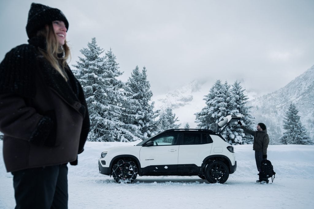 A woman stands near a white Jeep Avenger 4xe fitted with König XG Cross snow chains, looking at the snowy mountain landscape while a man closes the car trunk during snowfall.