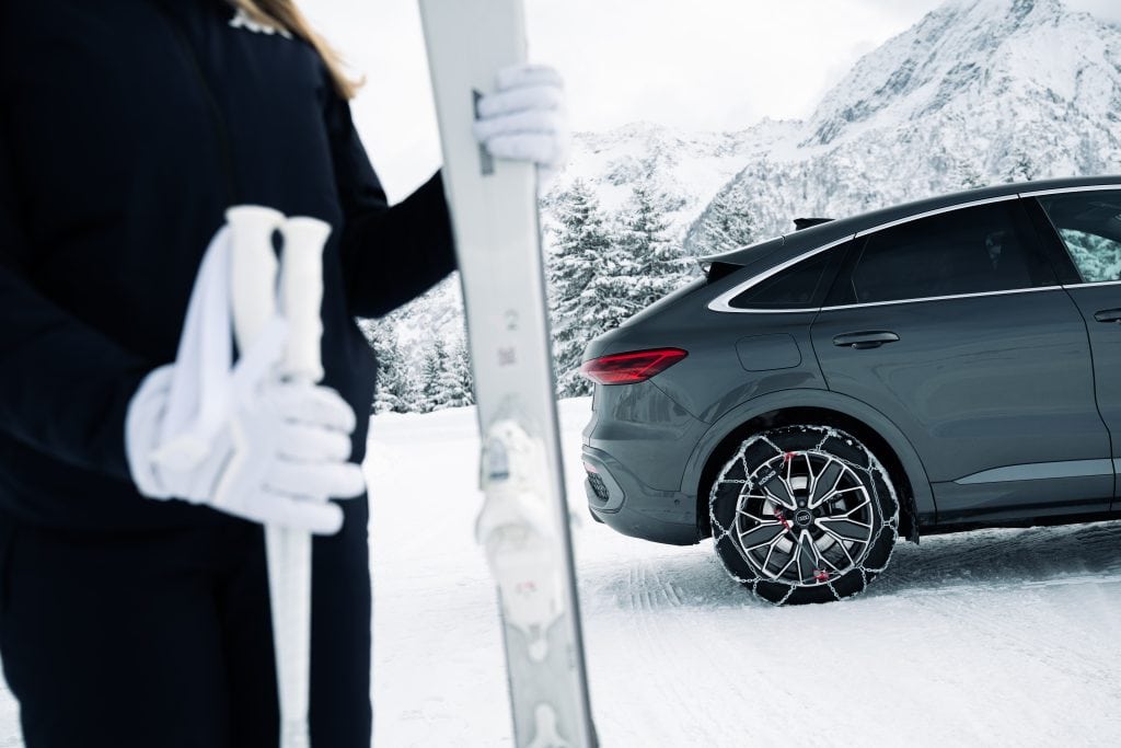 A woman with her skis stands beside a grey Audi Q5 fitted with König XG-12 Pro snow chains, parked in a snow-covered mountain area.