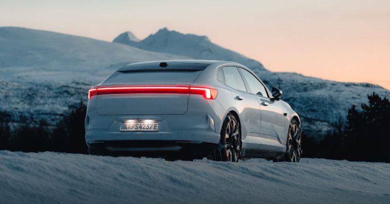 A white Polestar 4 fitted with König K-Summit MAX snow chains is parked on the snow at sunrise, with snow-covered mountains in the background.